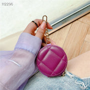 A person holds a small round quilted coin purse in a bright magenta pink color with a gold-tone keychain clip. The person is wearing a lavender long-sleeve shirt and denim shorts, sitting on a light-colored floor.