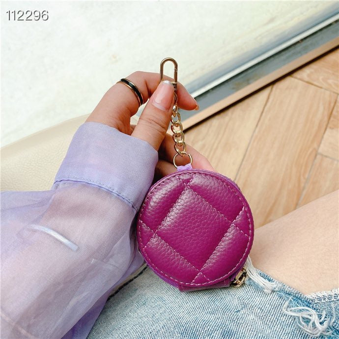 A person holds a small round quilted coin purse in a bright magenta pink color with a gold-tone keychain clip. The person is wearing a lavender long-sleeve shirt and denim shorts, sitting on a light-colored floor.