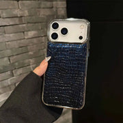 A hand holding a smartphone with a dark blue textured protective case featuring a glossy crocodile pattern and a clear bumper edge, set against a gray brick wall background.