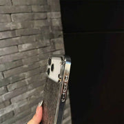 A close-up of a hand holding a smartphone with a clear, metallic, and textured protective case against a gray brick wall and black background.