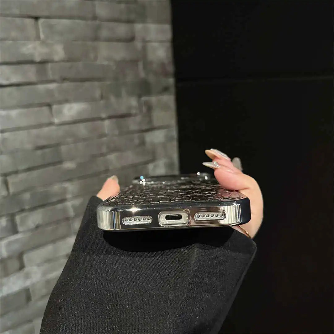 A close-up of a hand holding a smartphone case with a metallic silver keyboard design and a black textured back, positioned in front of a gray brick wall and a black background.