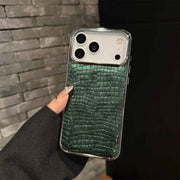 A hand holding a smartphone with a dark green crocodile-embossed leather back cover and a clear bumper frame, set against a dark gray brick wall background.