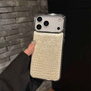 A hand holding a smartphone case with a light beige crocodile-embossed backplate and a clear bumper frame, shown against a dark gray brick wall background.