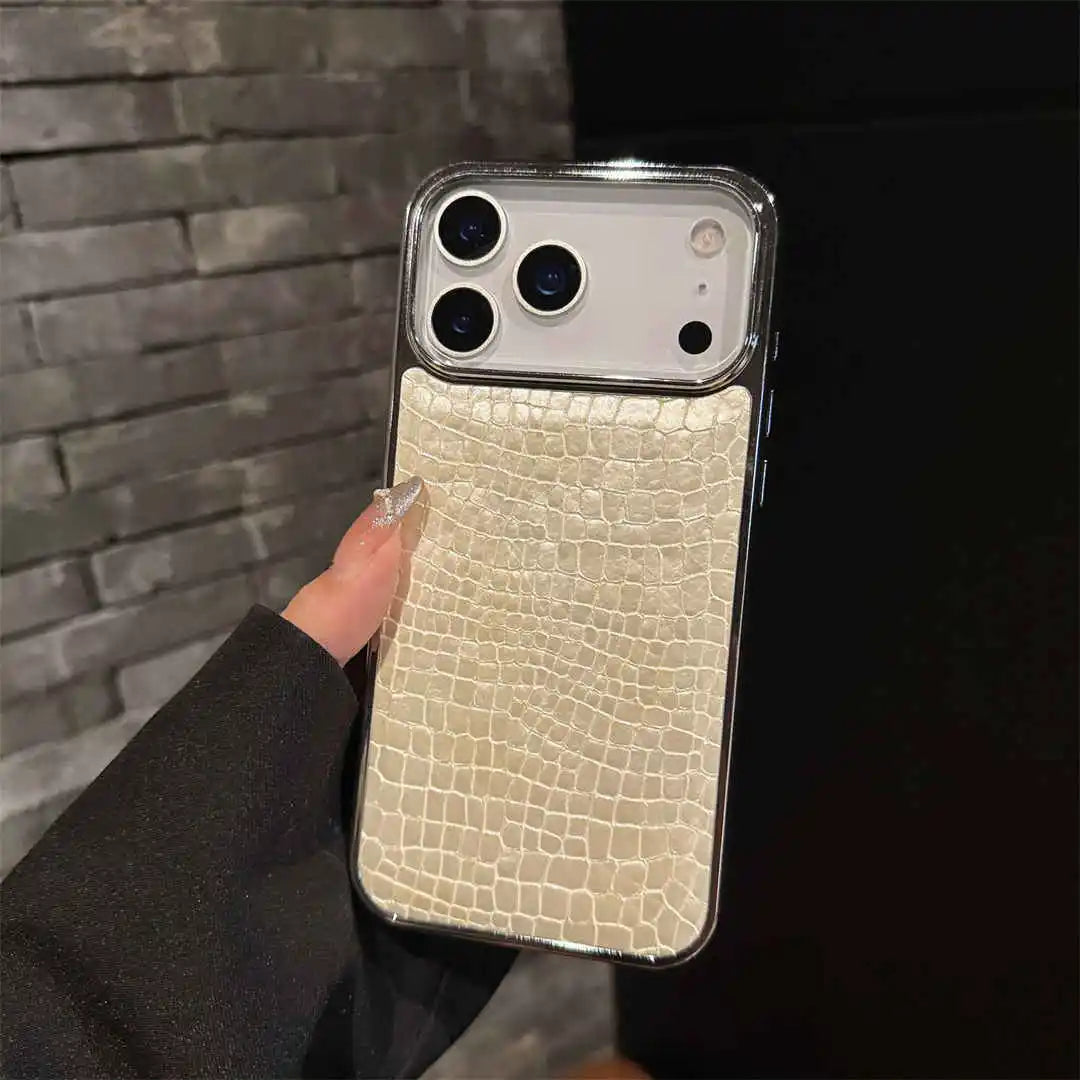 A hand holding a smartphone case with a light beige crocodile-embossed backplate and a clear bumper frame, shown against a dark gray brick wall background.