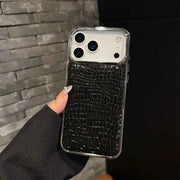 A hand with long manicured nails holds a smartphone case featuring a black crocodile-embossed texture on the back, with a clear transparent bumper edge, set against a dark gray brick wall background.
