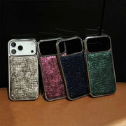 Four smartphone cases with a textured crocodile skin pattern and metallic trim, shown in silver, pink, navy, and green, lined up on a brown surface against a black background.