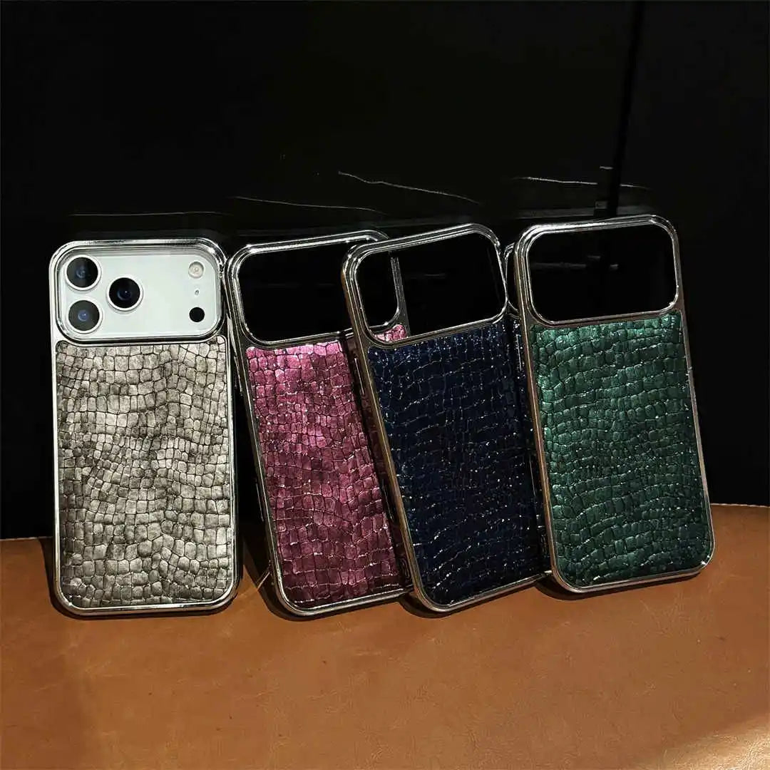 Four smartphone cases with a textured crocodile skin pattern and metallic trim, shown in silver, pink, navy, and green, lined up on a brown surface against a black background.