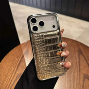A hand holding a smartphone case with a metallic gold and silver crocodile-embossed pattern on a wooden table.