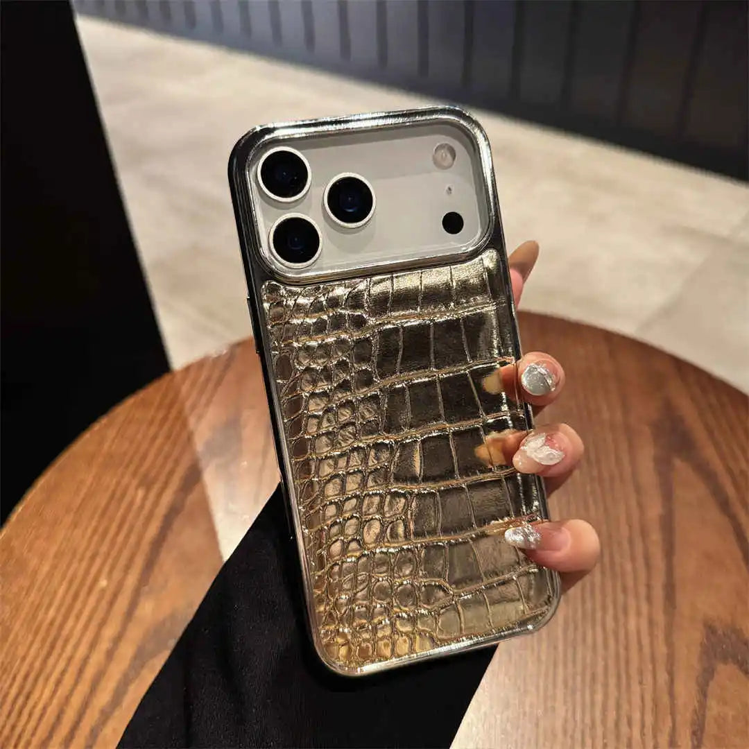 A hand holding a smartphone case with a metallic gold and silver crocodile-embossed pattern on a wooden table.
