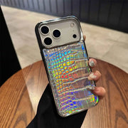 A hand holding a smartphone case with a silver metallic frame and an iridescent holographic crocodile-embossed back panel, shown over a wooden table.