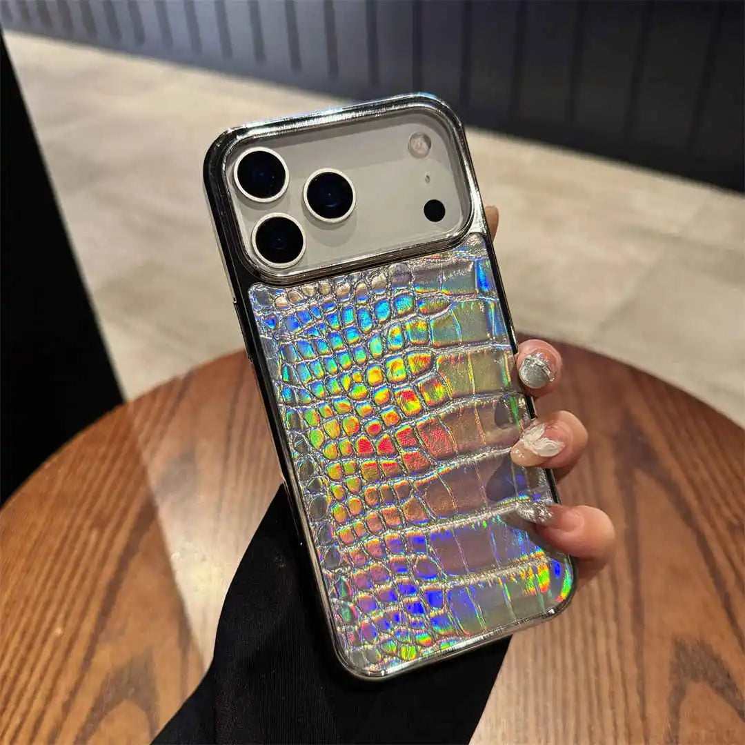 A hand holding a smartphone case with a silver metallic frame and an iridescent holographic crocodile-embossed back panel, shown over a wooden table.